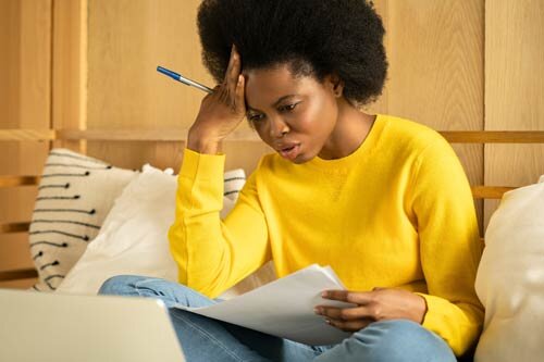 A black woman in a yellow T-shirt staring at a document in a worried state.